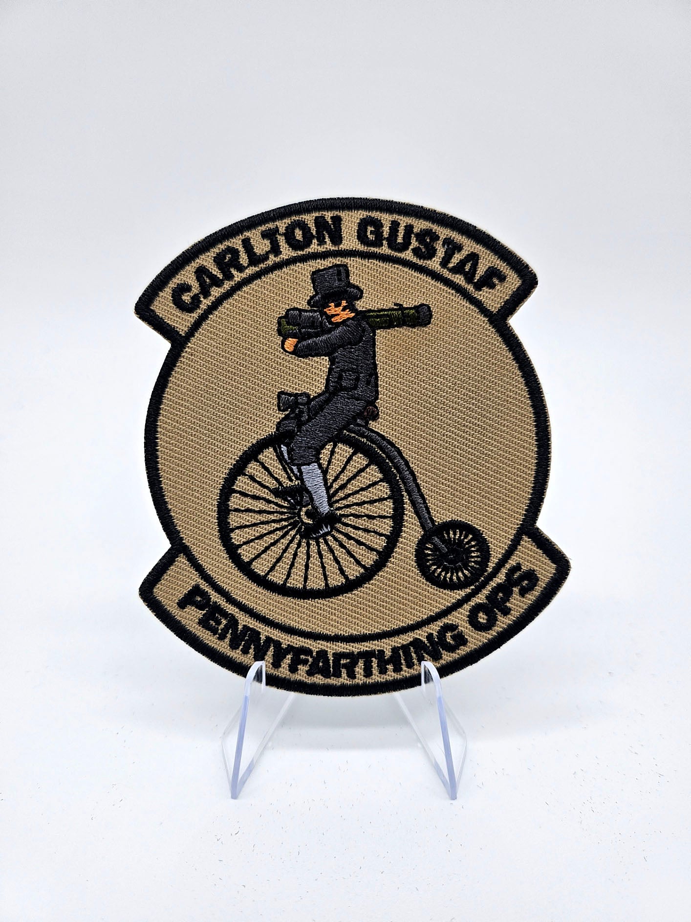 Patch with a person on a penny farthing bicycle and text 'Carlton Gustaf Pennyfarthing Ops' on a white. It features a fancy man on a Pennyfarthing holding a Carl Gustav shoulder mounted anti air weaponbackground