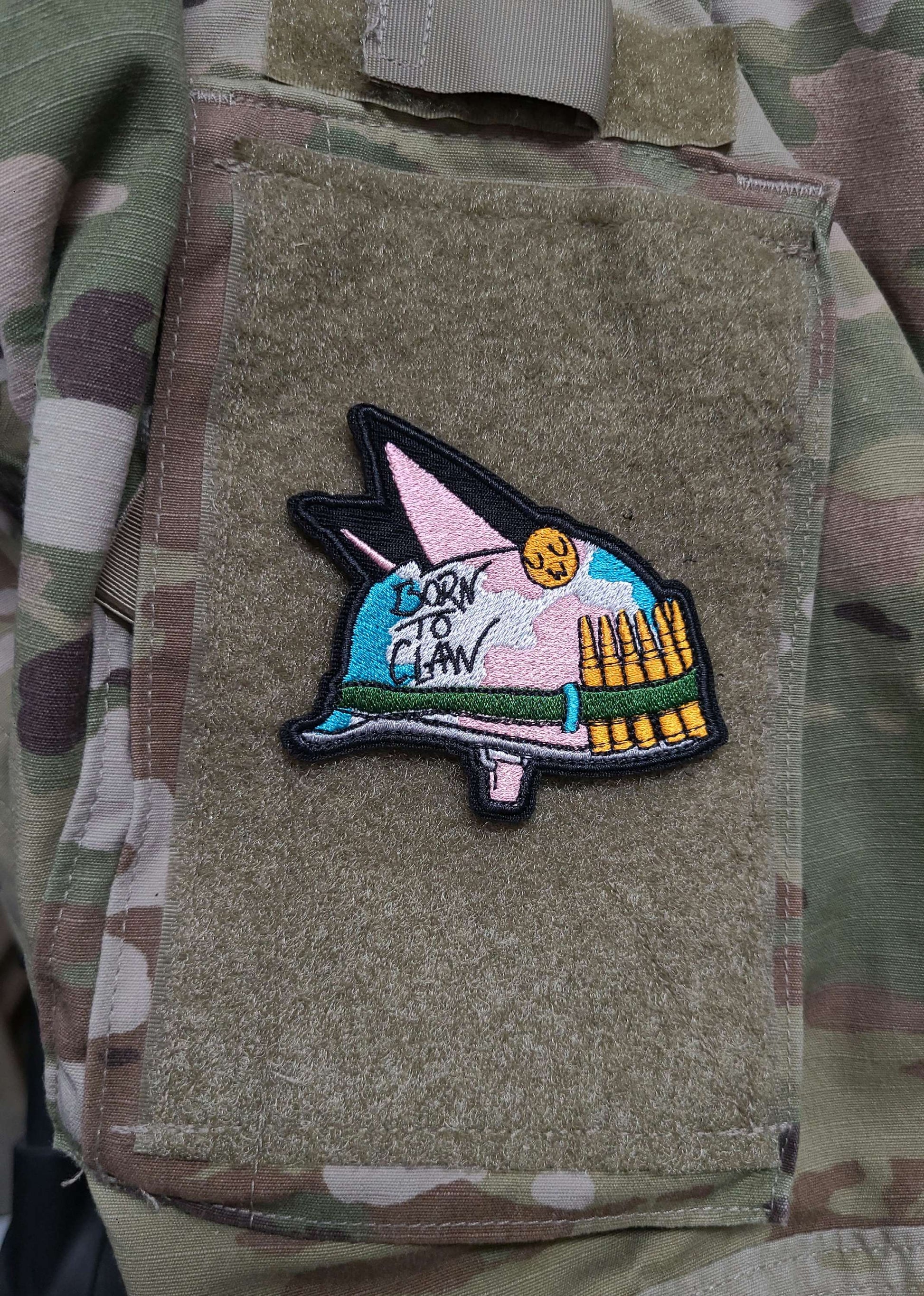 A velcro patch in the shape of an Army helmet with cat ears. The camoflague is in the pastel colors of the trans flag. Written on the side is Born to Claw