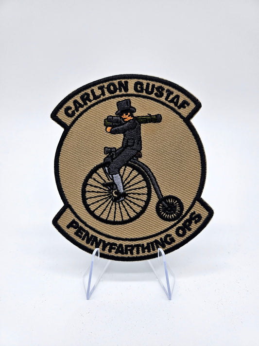 Patch with a person on a penny farthing bicycle and text 'Carlton Gustaf Pennyfarthing Ops' on a white. It features a fancy man on a Pennyfarthing holding a Carl Gustav shoulder mounted anti air weaponbackground