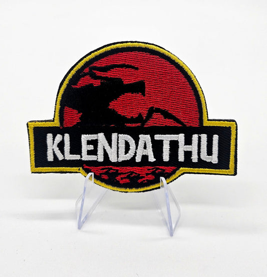 Embroidered patch with 'Klendathu' text and red and black design on a stand against a white background. It is an homage to the classic Jurassic Park logo, but with the bugs from Starship Troopers