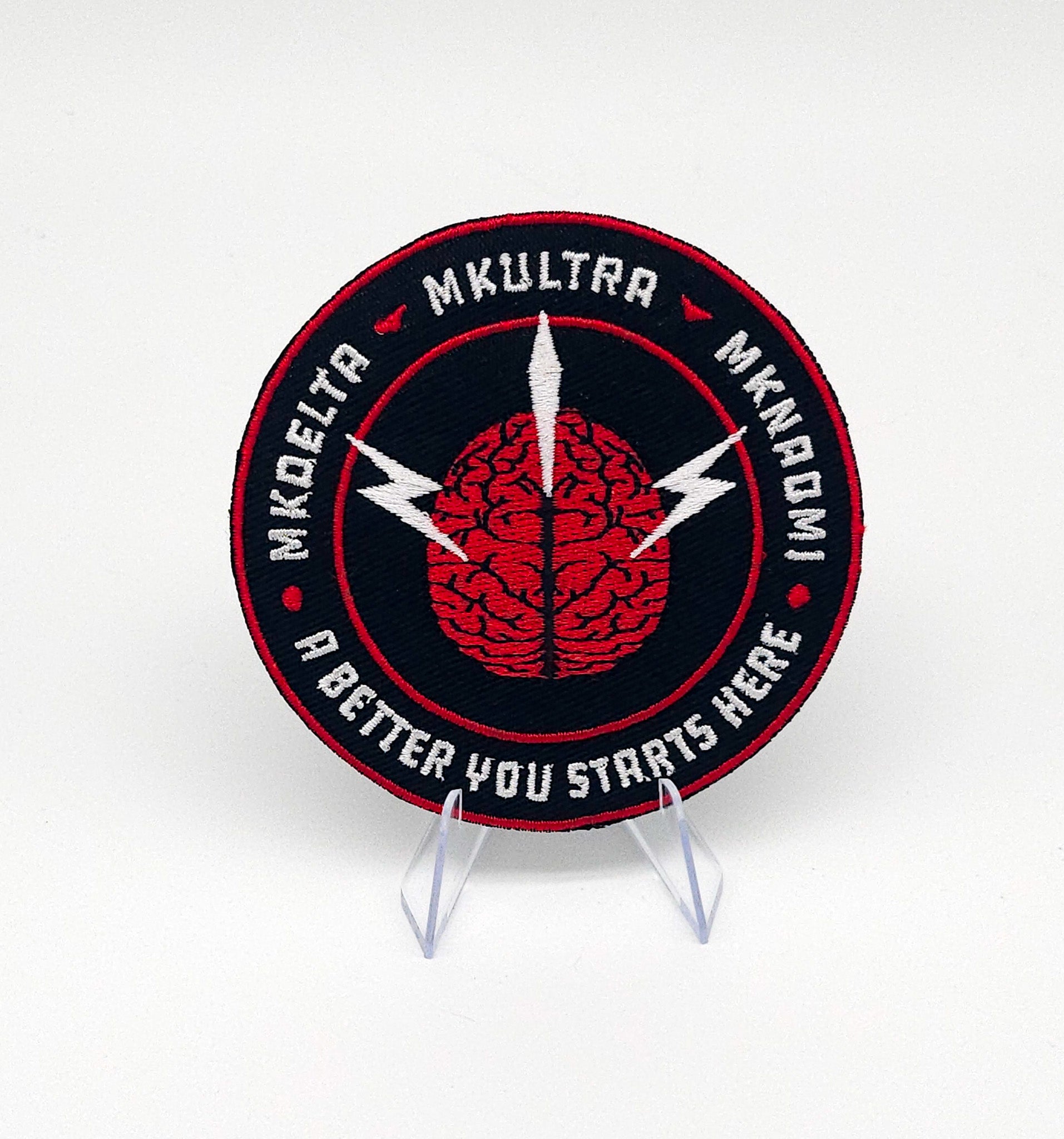 Circular patch with brain design and text on a stand against a white background. Inside the circle is MK DELTA MKULTRA and MKNAOMI with a brain in the middle. At the bottom it reads A better you starts herebackground