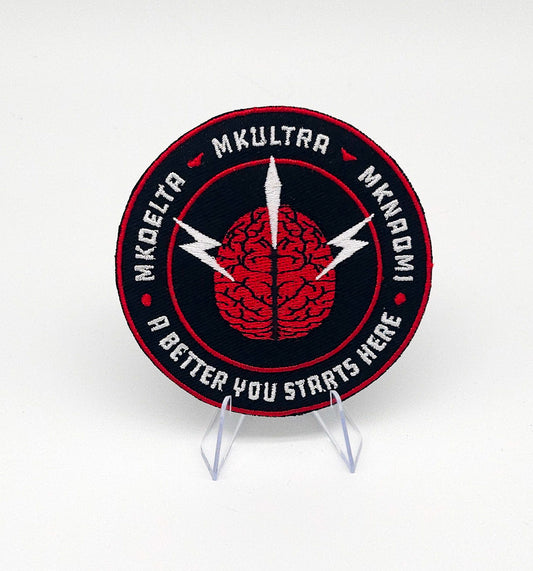 Circular patch with brain design and text on a stand against a white background. Inside the circle is MK DELTA MKULTRA and MKNAOMI with a brain in the middle. At the bottom it reads A better you starts herebackground