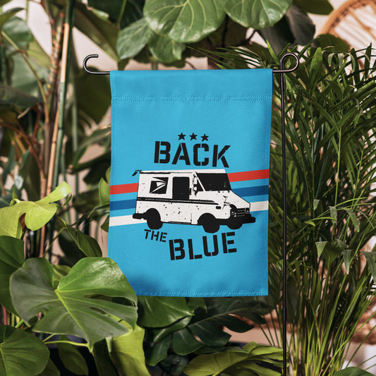 Blue garden flag with a truck graphic and text, hanging among green plants.